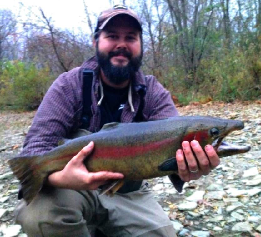 A man with a beard and in outdoor clothing holds a large fish with red and green coloring, standing on a rocky ground with trees in the background.